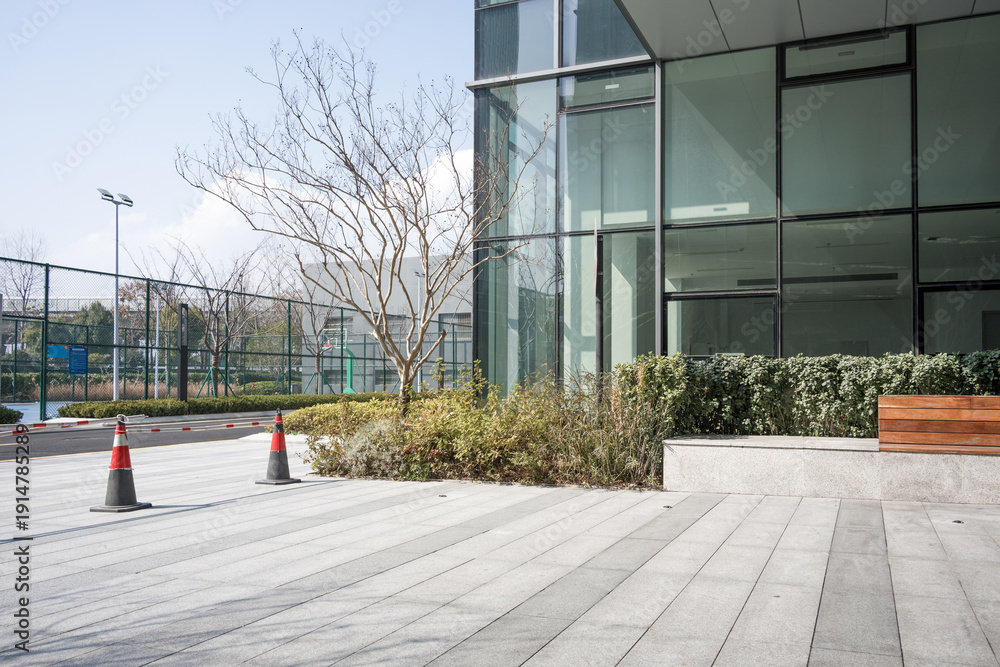 Obraz premium Modern glass building with empty paved plaza and landscaped greenery in daylight