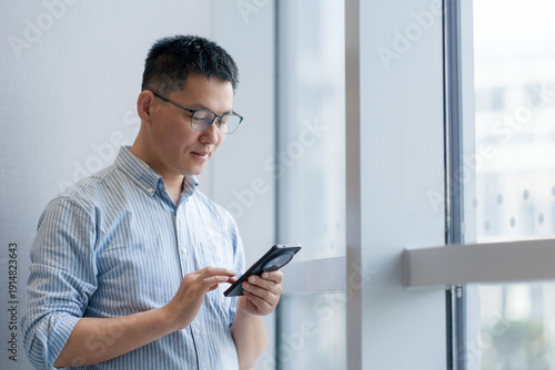 Canvas Print A middle-aged Asian man wearing glasses focuses intently on operating a smartpho