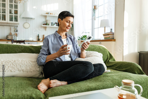 Relaxed pretty woman of 60s enjoying leisure time at home, sitting on green couch barefoot and in casual clothes drinking tea or coffee and using smartphone, chatting with chat bot