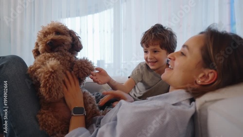 Mother and young child playing with a small dog on a cozy sofa at home. Warm family moment filled with joy, laughter, and bonding. Concept of family time, childhood happiness, pet love, and everyday