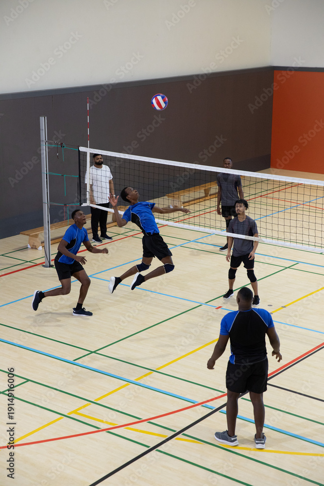 Fototapeta premium Opposing male volleyball teams leaping at net, spiking ball in gym with blue jerseys, knee-pads