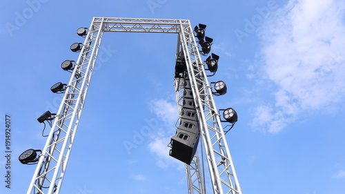 Speaker and lighting system on a metal frame. Line array speakers hanging from a pole beside an outdoor concert stage on a blue sky background with text copy space with selective focus.
