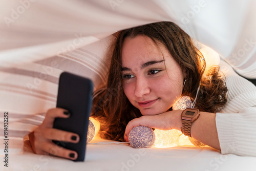 Smiling teenage girl with chain of lights using cell phone underneath bedcover