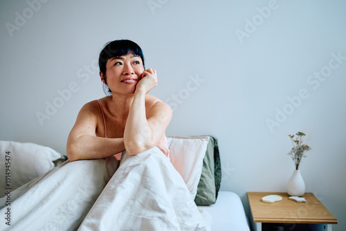 Tableau sur toile Asian woman sitting up in cozy bed, calmly contemplating morning thoughts with a