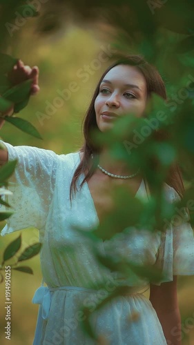 Young woman in white dress and pearl necklace peeking through green leaves in soft sunlight