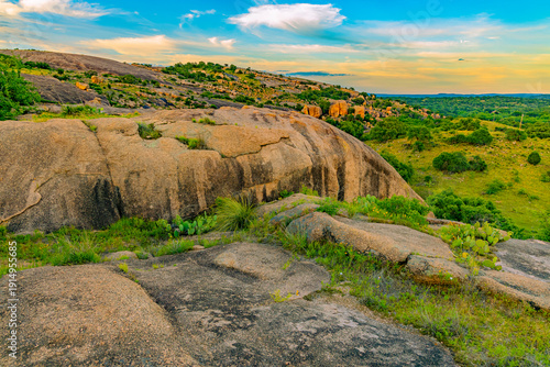 Slika na platnu Expansive view from atop the pink granite dome, showcasing layered rock formations, lush green hills, and a vibrant sunset sky in Enchanted Rock State Natural Area, Texas