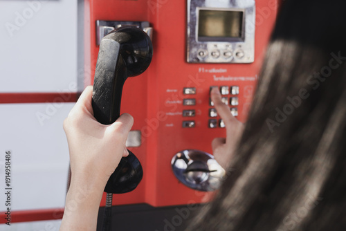 A close-up of vintage black telephone handset, with a hand pressing buttons on classic red public telephone. Concepts of retro communications and network technology.