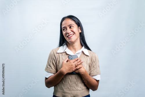 Fotografija A cheerful young Asian woman in a beige vest holding her mobile phone close to her heart and smiling joyfully, isolated on a white background