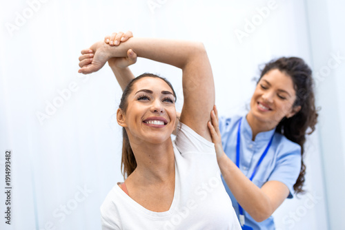 A smiling patient performs shoulder stretching exercises assisted by a female physiotherapist. Professional rehabilitation and physical therapy for arm mobility and injury recovery in a bright office.