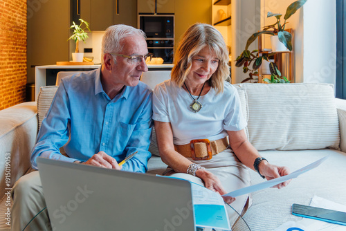 Photography Senior couple on sofa at home reviewing bills and documents, using a laptop to d