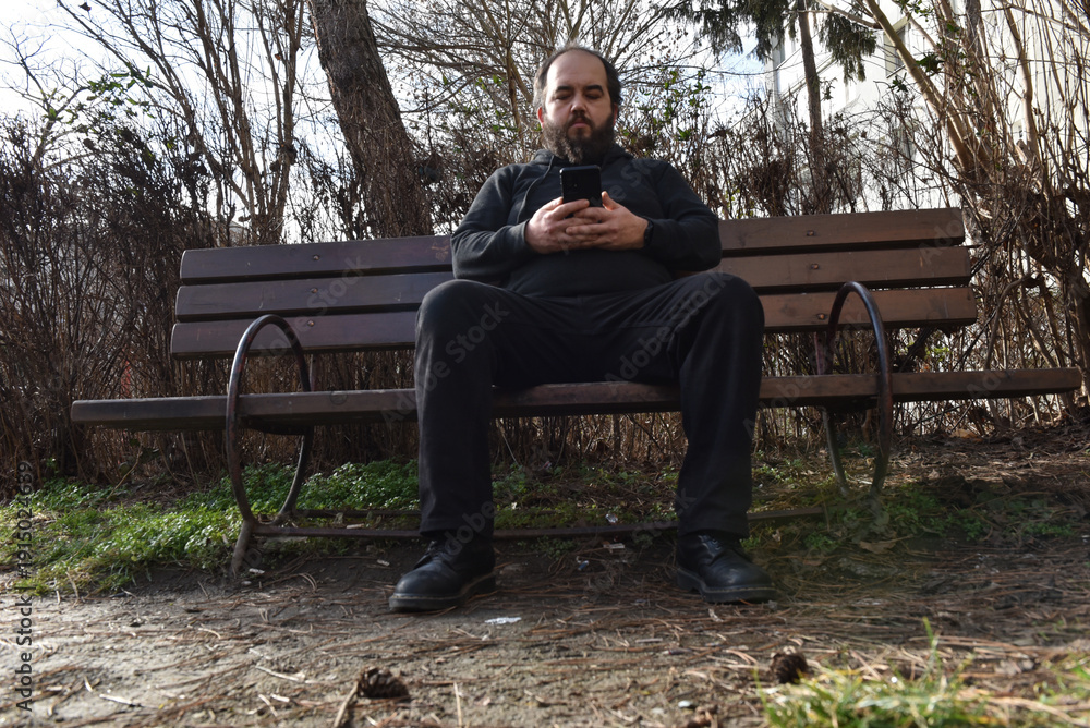 Fototapeta premium Bearded man in black casual clothing sitting on a wooden park bench while looking at his smartphone