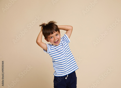 Frontal portrait of a happy little boy in a striped t-shirt, posing in the studio, making faces, having fun, on a beige background.