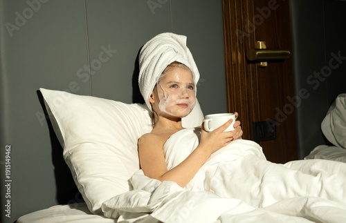 Little girl with towel on her head lying in bed with maca on her face. Skin care, moisturizing masks