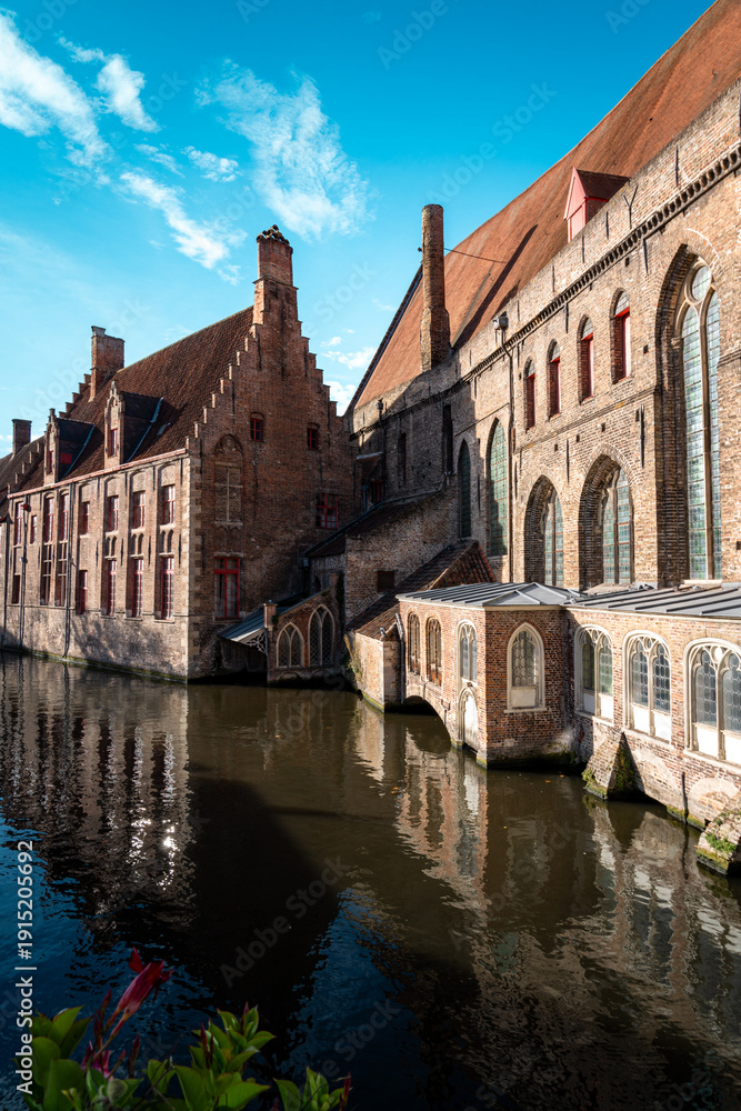 Obraz premium portrait shot of buildings over canal river from bridge in bruges in belgium with orange gothic details on the stone and cloudy clear blue skies above in autumn
