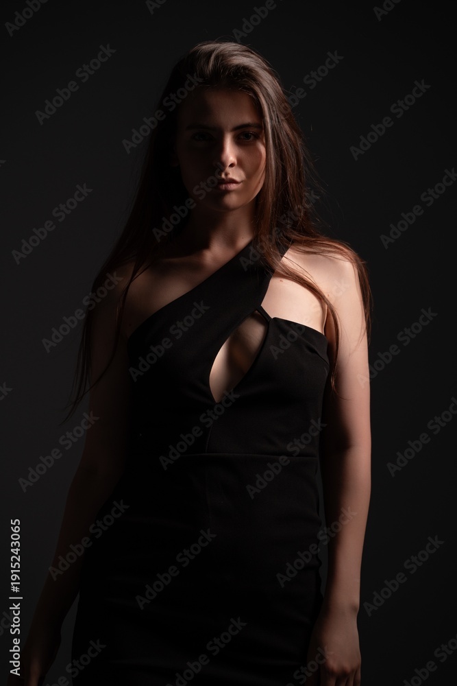 Naklejka premium Woman posing in elegant black dress with cutout detail, capturing beauty, fashion, and mysterious mood through low-key studio lighting