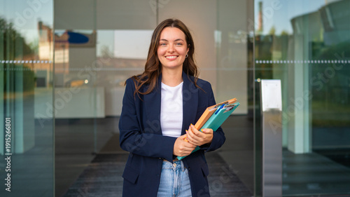 Confident young businesswoman holding folders smiling at office entrance