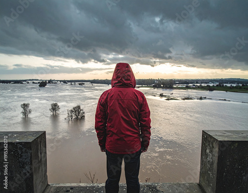 A man in a red jacket stands on a ledge overlooking a flooded area. The sky is cloudy and the water is murky