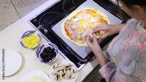Adorable young girl preparing homemade pizza by placing ingredients on fresh dough. 