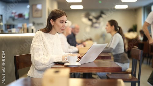  In cozy cafe hall, female freelance works with laptop and drinks coffee. Remote work from anywhere in world. Internet cafe, catering and workshop zone area