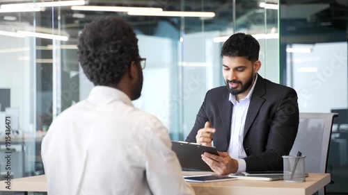 HR manager sitting at office desk talking to candidate during job interview and writing notes on clipboard. Employer evaluating applicant, listening, assessing experience in corporate environment.