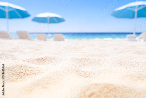 Close-up of clean white sand in the foreground with blurred blue sun umbrellas and lounge chairs on a sunny beach background. Concept of summer vacation and luxury resort relaxation