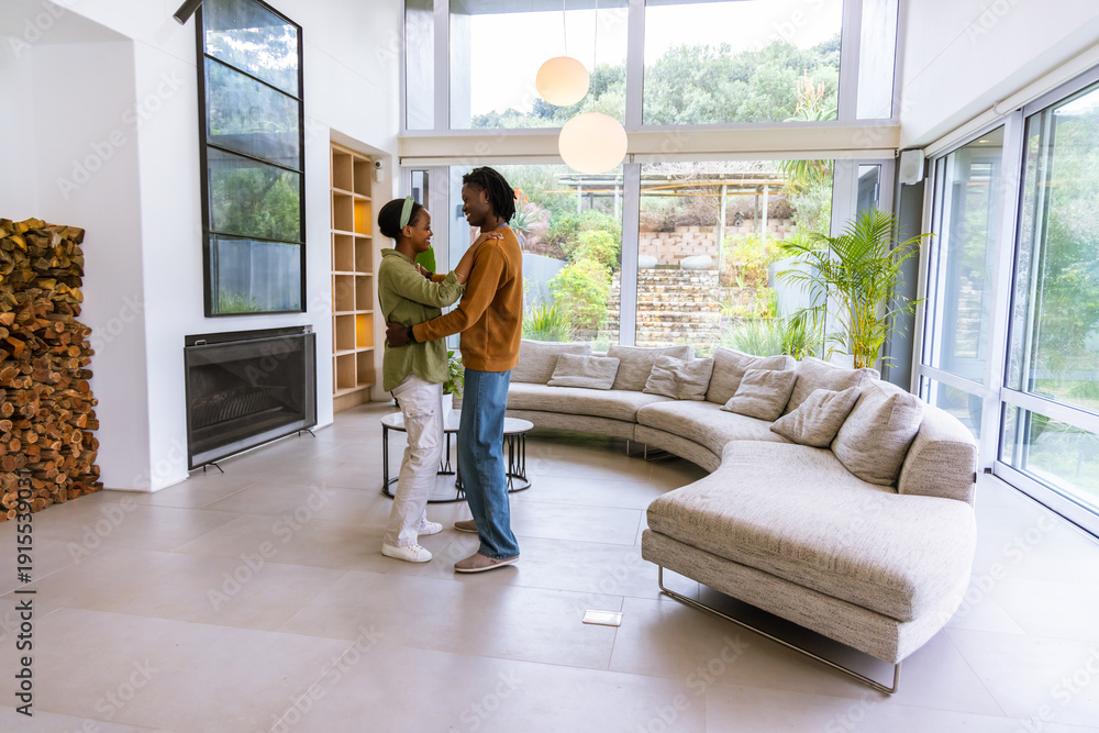 Naklejka premium African American couple embracing and standing in modern living room near fireplace and sofa