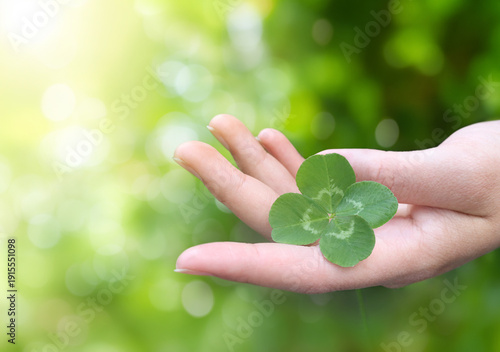 Schilderij op canvas A woman's hand gently holds a vibrant four-leaf clover against a soft green bokeh background in warm sunlight