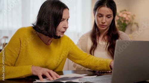 Experienced Caucasian woman pressing laptop keys at table. Beautiful female guiding and watching screen attentively. Papers lying nearby during lesson. Learning digital skills at home kitchen.