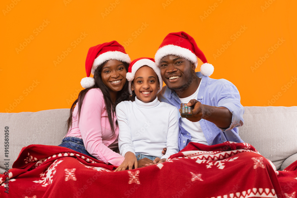 Fototapeta premium A family sits on a couch wrapped in a blanket. They smile and pose for the camera. All three family members wear red Santa hats. They are in a cozy room with an orange wall.