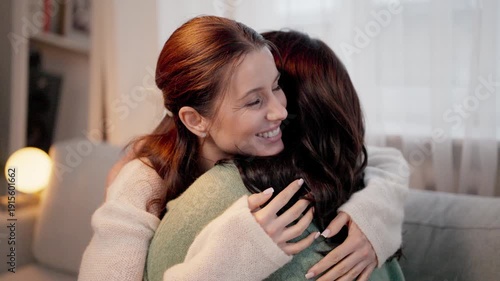 Pretty woman with long hair warmly embracing dark haired female while sitting on sofa. Charming daughter hugging her beloved mother with sincere smile. Expressing tenderness affection.