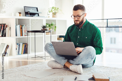 Quadro su tela Man Using Laptop Computer Sitting On Floor Wearing Glasses, Working Online Browsing Internet And Typing At Home