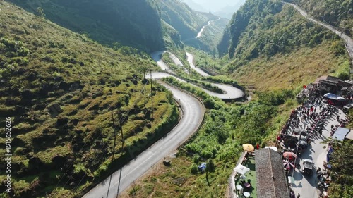 Aerial view of the Doc Tham Ma Pass, Dong Van, Ha Giang, Vietnam. Cars and motorbikes navigate the winding road, while tourists gather at a viewpoint to enjoy the scenery.