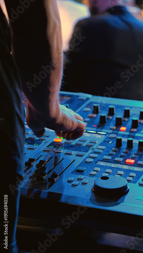 Close-up of a sound engineer's hand on a mixing board, adjusting faders. The console is illuminated by cool blue light in a dark studio concert venue. Professional, focused, creative atmosphere