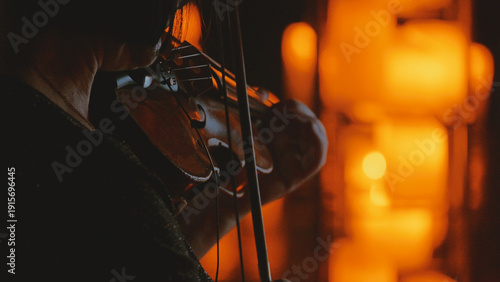 Close-up of a violinist performing in a dark, intimate setting. The musician is silhouetted, lit by a warm, golden glow from blurred candlelight, creating a moody and atmospheric bokeh effect