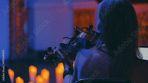 Over-the-shoulder shot of a woman playing the violin at an intimate concert. Dramatic blue and warm orange light from candles creates a moody, atmospheric and cinematic feel