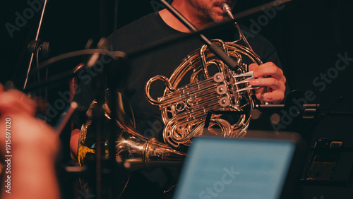 A musician plays a brass French horn in a symphony orchestra. Warm, low-key lighting highlights the instrument against a dark background, creating a moody, artistic atmosphere for a live performance