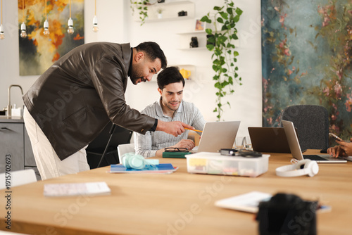 Two men collaborate on a project at a wooden table, one pointing at a laptop screen with a pencil, surrounded by office supplies and modern decor.