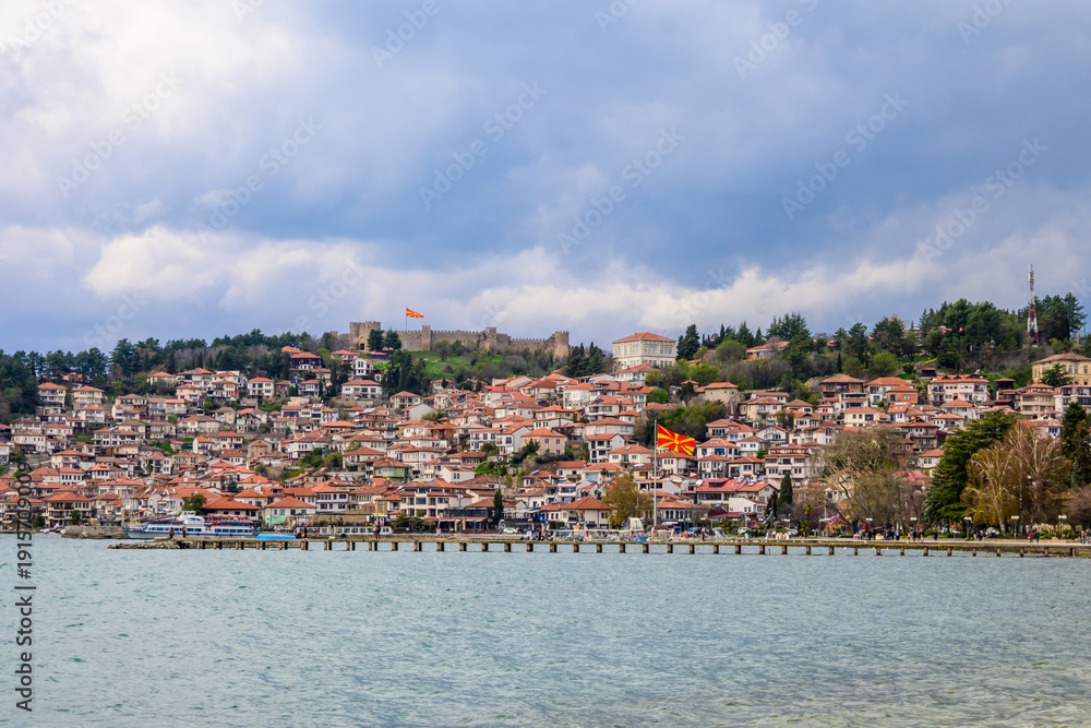 Obraz premium Historic Ohrid town with traditional red-roofed houses climbing a hillside, crowned by an ancient fortress, overlooking a vast lake under a cloudy atmosphere.