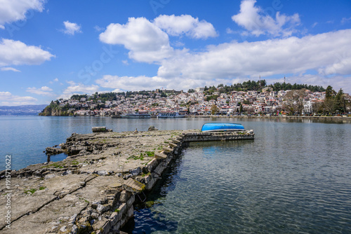 A textured stone pier and upturned blue boat extend into clear lake waters, framed by the distant terraced houses of historic Ohrid city and its fortress.
