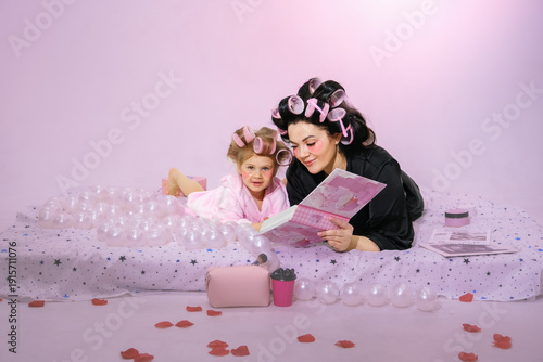 Moscow, Russia, 08.02.2026 Woman and girl with hair curlers read a book on a bed, skin care beauty concept. For Mothers Day greeting card.