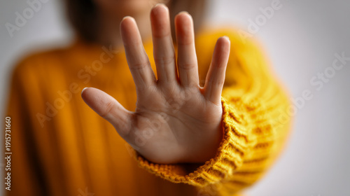 Person wearing a cozy yellow sweater with an outstretched hand signaling stop or refusal against a soft blurred background in a gesture of boundary setting or protec
