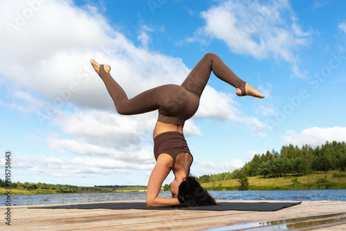 A woman performs a variation of the Sirsasana exercise, an inverted pose, a stand on her elbows with her legs extended, and practices yoga on the shore of a lake