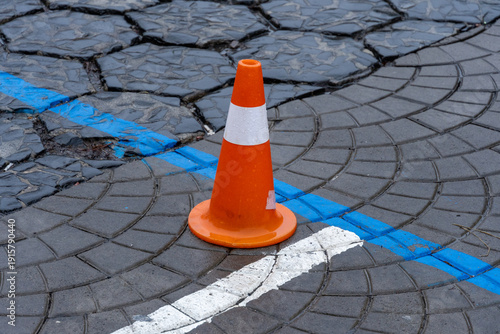 An orange traffic cone stands prominently on a patterned road, contrasting against the cracked surface and blue markings during the day