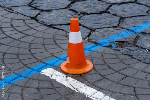 An orange traffic cone rests on cracked, uneven pavement with blue and white lines, indicating a parking area or construction zone