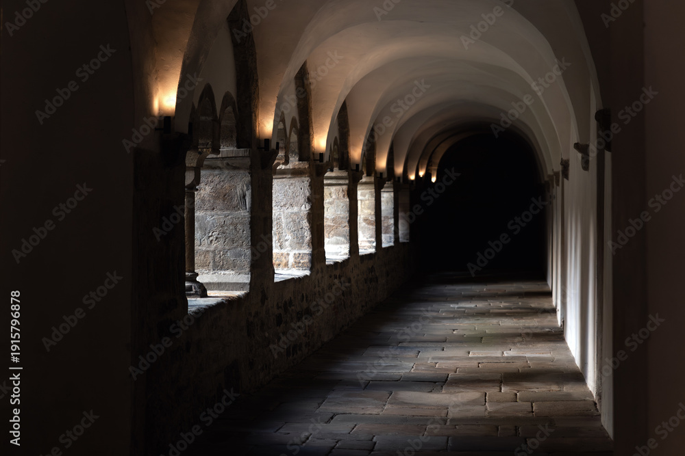 Fototapeta premium Medieval cloister corridor stone arches vaulted ceiling darkness illuminated warm sconces textured masonry wall. Perspective shadow silence create sacred atmosphere inside historic European monastery