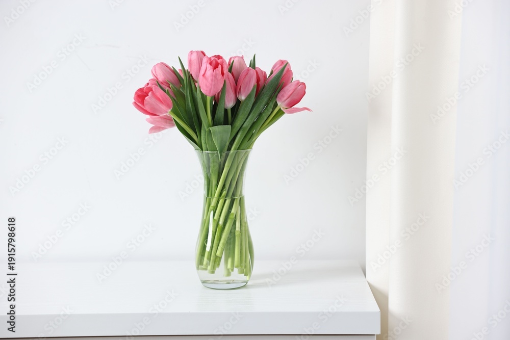 Fototapeta premium Bouquet of pink tulips in vase on chest of drawers near white wall indoors