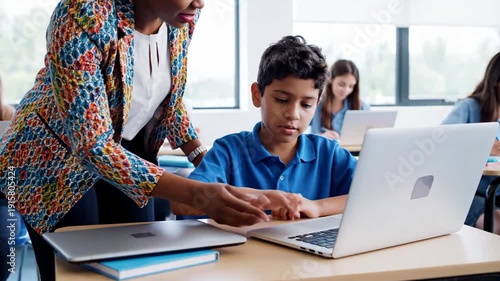 Teacher assisting student with laptop computer in modern classroom setting with other students working on their own devices.