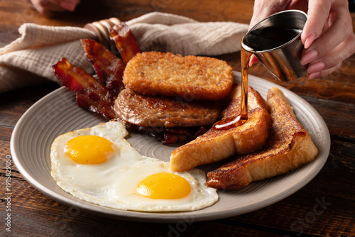 Obraz na płótnie Close-up rustic shot of a woman pouring syrup on French toast on a plate with eggs, bacon, sausage, and hash browns