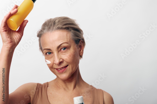Φωτογραφία Mature woman with short grey hair applies sunscreen cream on face holding bottle, smiling gently on white background