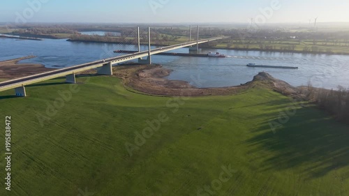 Wallpaper Mural Aerial view of a cable stayed bridge over a wide river, with two cargo vessels below and green fields, trees, and distant wind turbines forming a calm rural backdrop. Torontodigital.ca